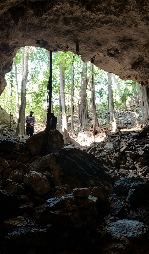 Entrada de una cueva con roca oscura en primer plano y árboles al fondo; una persona al centro